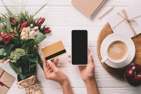 Обрезанный снимок женщины, держащей кредитную карту и смартфон с чистым экраном на столе с букетом цветов и чашкой кофе — стоковое фото