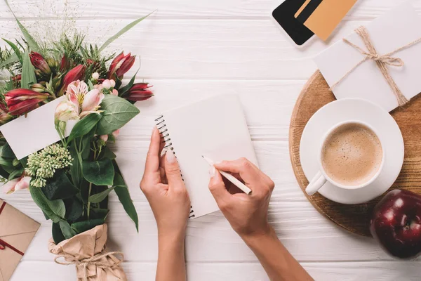 Vista parcial de la mujer haciendo notas en el cuaderno en la mesa con ramo de flores y taza de café - foto de stock