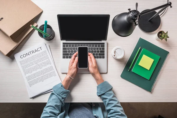 Imagen recortada de hombre de negocios utilizando el teléfono inteligente en la mesa con el ordenador portátil, contrato y taza de café desechable - foto de stock