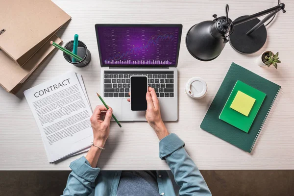 Ausgeschnittenes Bild eines Geschäftsmannes mit Smartphone am Tisch mit Vertrag, Schreibwaren und Laptop mit Grafik auf dem Bildschirm — Stockfoto