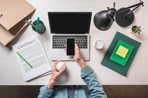 Imagen recortada de hombre de negocios con bola para el béisbol utilizando el teléfono inteligente en la mesa con el ordenador portátil y contrato - foto de stock