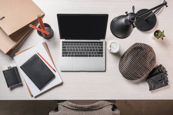 Erhöhte Sicht auf den Arbeitsplatz mit Fläschchen, Kaffee, Mütze, Schreibwaren und Laptop auf dem Tisch — Stockfoto