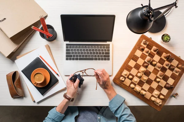 Обрізане зображення чоловічого фрілансера, що протирає окуляри за робочим столом з ноутбуком, шахівницею та кавою — стокове фото