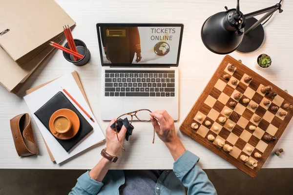 Imagen recortada de hombre freelancer limpiando anteojos en la mesa con tablero de ajedrez, café y portátil con entradas en línea en la pantalla - foto de stock