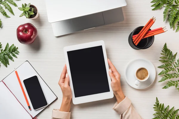 Vista parcial del freelancer femenino sosteniendo tableta digital con pantalla en blanco en la mesa con gadgets y café rodeado de hojas de helecho - foto de stock