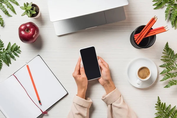 Visão parcial de freelancer feminino usando smartphone com tela em branco à mesa com papelaria, laptop e café cercado por folhas de samambaia — Fotografia de Stock