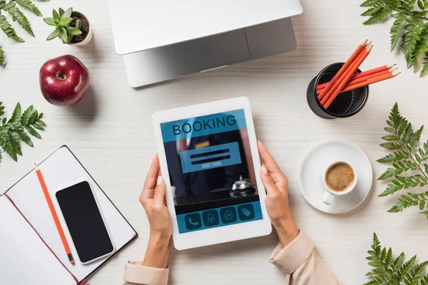 Imagen recortada de freelancer femenina sosteniendo tableta digital con reserva en pantalla en la mesa con gadgets y café rodeado de hojas de helecho - foto de stock