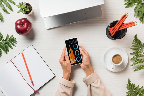 Vista parziale di freelance utilizzando smartphone con grafici e grafici sullo schermo a tavola con computer portatile, caffè, libro di testo circondato da rami con foglie verdi — Stock Photo