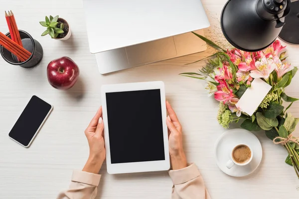 Частичный вид женщины-фрилансера, держащей цифровой планшет с чистым экраном за столом с чашкой кофе, гаджетами и цветами с поздравительной открыткой — стоковое фото