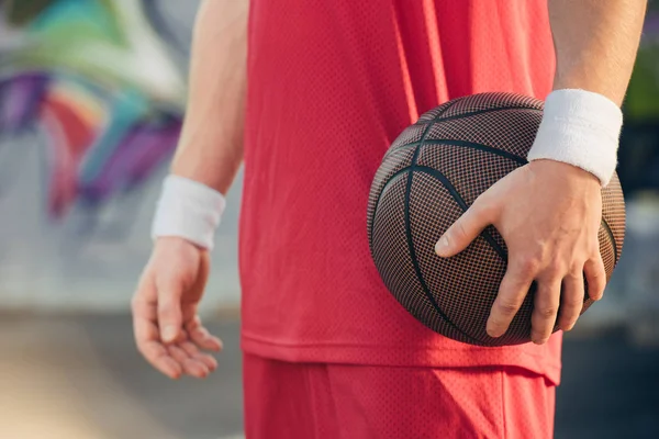 Cropped image of basketball player in red sportswear holding basketball ball on street — Stock Photo