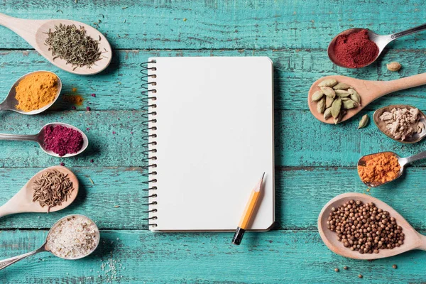 Top view of blank notebook with pencil and spoons with various spices on turquoise wooden surface — Stock Photo