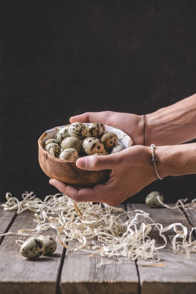 Cropped shot of person holding bowl with quail eggs above wooden table with shavings — Stock Photo