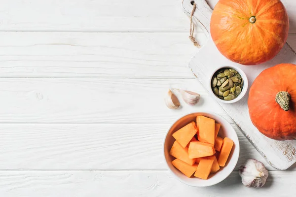 Elevated view of pumpkin pieces in bowl, pumpkin seeds and garlic on table — Stock Photo