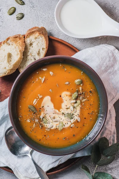 Elevated view of pumpkin cream soup with seeds in bowl and bread on wooden board on table — Stock Photo