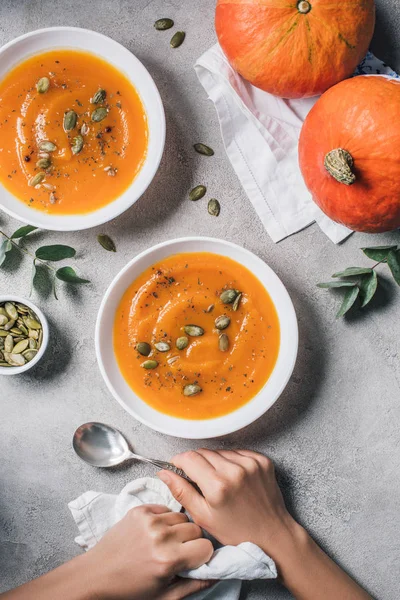 Partial view of woman sitting at table with pumpkins and plates with pumpkin soup and seeds — Stock Photo