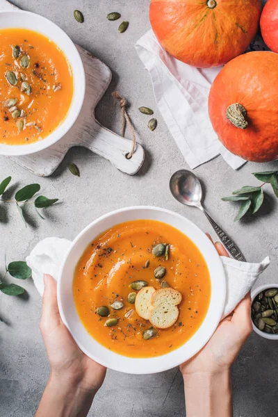 Immagine ritagliata di donna che tiene piatto con zuppa di crema di zucca fatta in casa con semi e fette biscottate sul tavolo — Foto stock