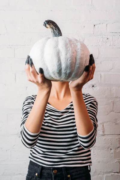 Mujer con camisa a rayas sosteniendo calabaza de halloween pintada de blanco delante de la cabeza - foto de stock