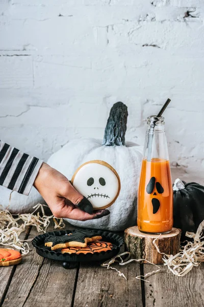 Cropped shot of woman holding halloween cookie near pumpkins and juice on wooden surface — Stock Photo