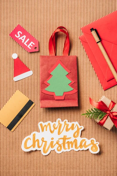 Top view of red shopping bag, decorated gift box, credit card and merry christmas lettering on surface — Stock Photo