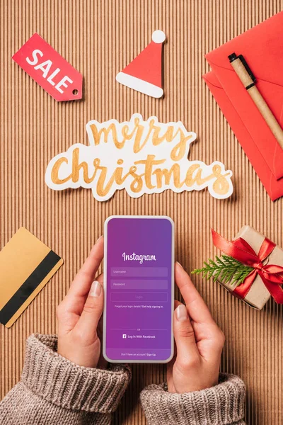 Partial view of woman holding smartphone with instagram on screen over surface with sale sign, credit card and merry christmas lettering — Stock Photo