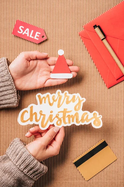 Cropped image of woman holding merry christmas lettering and santa hat over surface with credit card and sale — Stock Photo