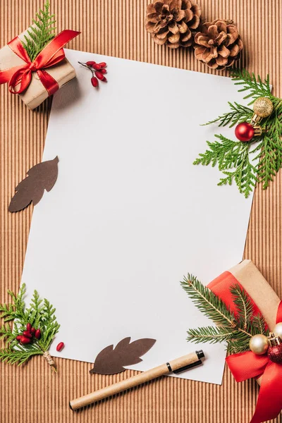 Vista elevada de papel vacío, conos siempreverdes, cajas de regalo decoradas con adornos de Navidad en la superficie - foto de stock