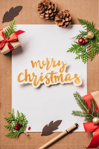 Vista superior de papel vacío, conos siempreverdes, cajas de regalo decoradas y letras Feliz Navidad en la superficie - foto de stock