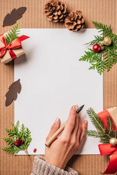 Image recadrée de femme avec stylo à la surface avec du papier vide et décoré boîtes cadeaux de Noël avec des boules et des branches à feuilles persistantes — Photo de stock