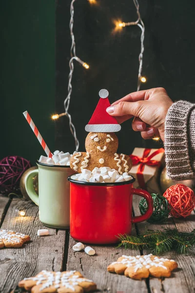 Mujer sosteniendo hombre de jengibre de Navidad en cacao con malvaviscos en mesa de madera con guirnalda ligera - foto de stock