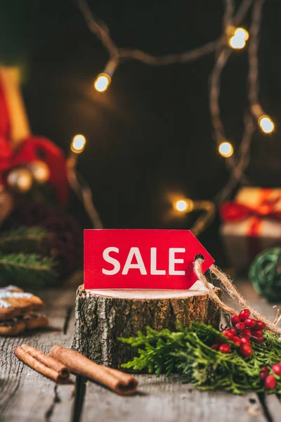 Red christmas sale tag on wooden stump with cinnamon sticks and light garland — Stock Photo