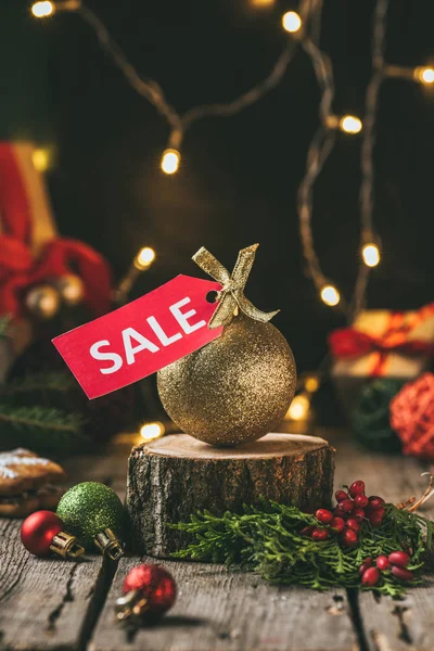 Golden christmas ball with red sale tag on wooden stump with light garland — Stock Photo