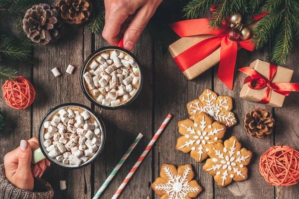 Cropped view of couple holding cocoa cups with marshmallows and gingerbread on wooden background with christmas decor — Stock Photo