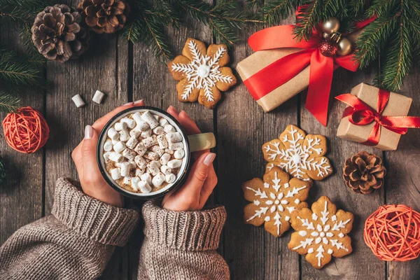 Vista recortada de la mujer sosteniendo taza de cacao con malvaviscos y pan de jengibre sobre fondo de madera con regalos de Navidad - foto de stock