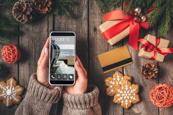 Vista recortada de la mujer usando el teléfono inteligente con la aplicación del boleto en el fondo de madera con tarjeta de crédito y regalos de Navidad - foto de stock