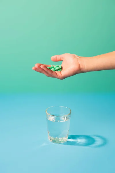 Recortado tiro de la persona sosteniendo píldoras por encima del vaso de agua en verde - foto de stock