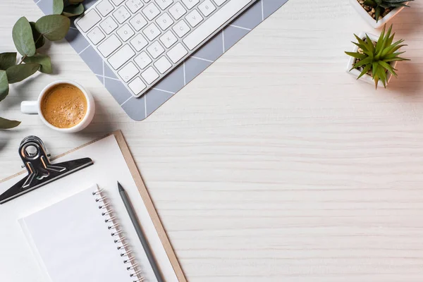 Vista superior del teclado de la computadora, libro de texto vacío, taza de café y plantas en maceta en la mesa en la oficina - foto de stock