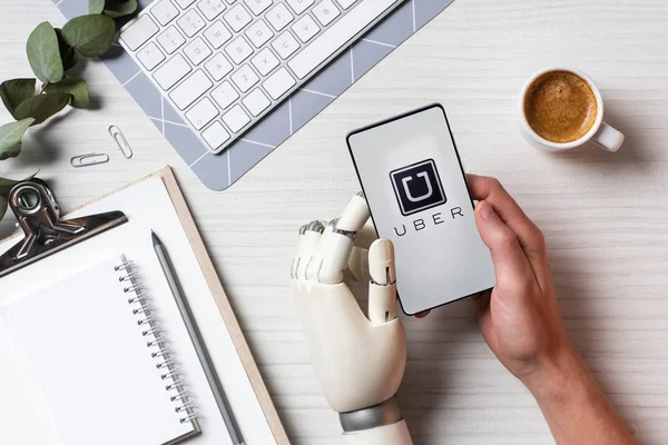 Vista parcial del hombre de negocios con brazo de prótesis usando smartphone con uber en pantalla en la mesa con taza de café en la oficina - foto de stock