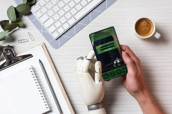 Обрезанный образ бизнесмена с протезом руки с помощью смартфона с бронированием на экране за столом с чашкой кофе в офисе — стоковое фото