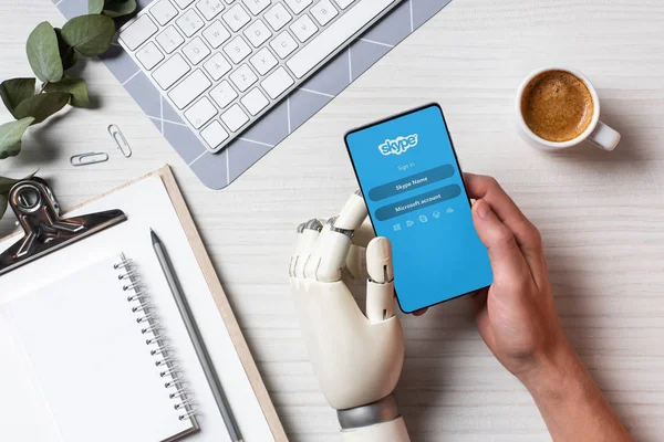 Vista parcial del hombre de negocios con brazo de prótesis usando teléfono inteligente con Skype en la pantalla en la mesa con taza de café en la oficina - foto de stock