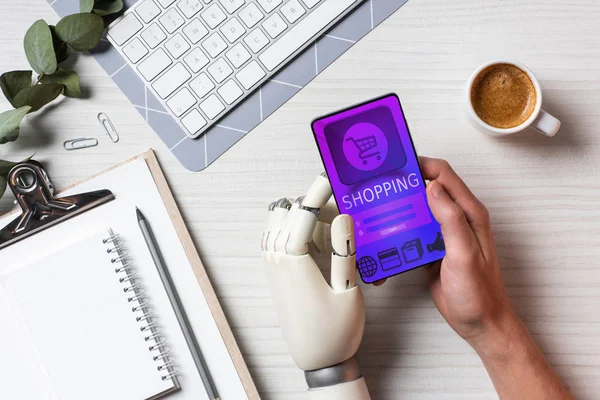 Partial view of businessman with prosthesis arm using smartphone with shopping on screen at table with coffee cup in office — Stock Photo