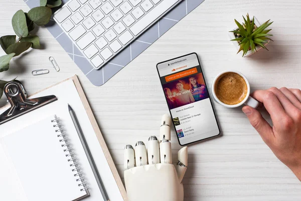 Imagen recortada de hombre de negocios con brazo de prótesis utilizando smartphone con soundcloud en pantalla en la mesa con taza de café en la oficina - foto de stock