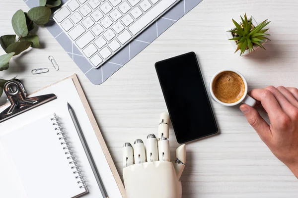 Частковий вид на бізнесмена з протезом рукою за допомогою смартфона з порожнім екраном за столом з чашкою кави в офісі — стокове фото