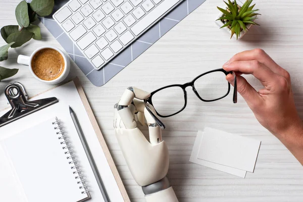 Image recadrée de l'homme d'affaires avec cyborg main tenant des lunettes à table avec cartes de visite vides et clavier d'ordinateur dans le bureau — Photo de stock