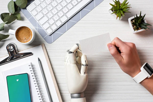 Partial view of businessman with cyborg hand and smartwatch holding blank visit card at table with smartphone with youtube on screen in office — Stock Photo