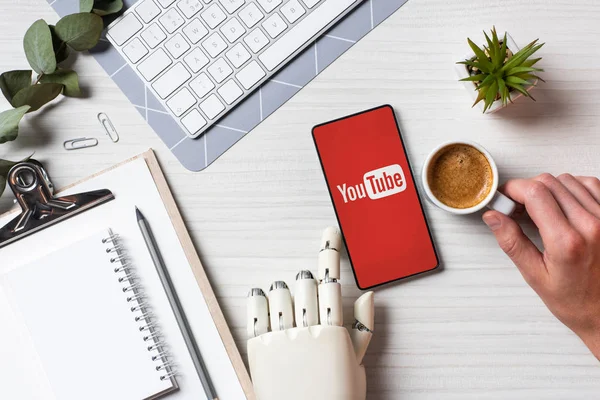 Cropped image of businessman with prosthesis arm using smartphone with youtube on screen at table with coffee cup in office — Stock Photo