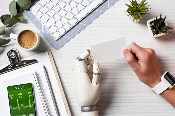 Cropped image of businessman with cyborg hand and smartwatch holding blank visit card at table with smartphone with medical application on screen in office — Stock Photo