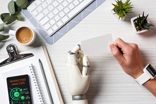 Partial view of businessman with cyborg hand and smartwatch holding blank visit card at table with smartphone with marketing analysis on screen in office — Stock Photo