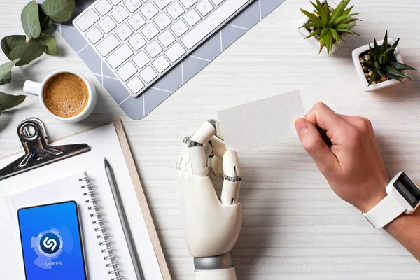 Обітнутого зображення бізнесмен з кіборг руку і smartwatch холдингу пусті візитна картка столом з смартфон з shazam на екрані в офісі — стокове фото