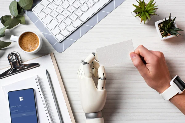 Cropped image of businessman with cyborg hand and smartwatch holding blank visit card at table with smartphone with facebook on screen in office — Stock Photo