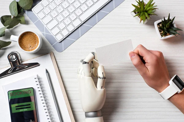 Cropped image of businessman with cyborg hand and smartwatch holding blank visit card at table with smartphone with booking on screen in office — Stock Photo